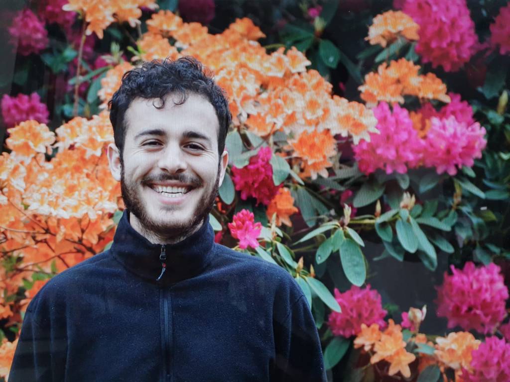 Life coach Yusef Sanei smiling in a navy fleece in front of orange and pink rhododendrons in bloom