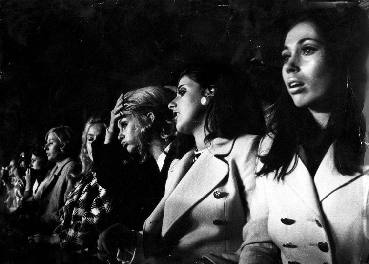 Black and white photo of a row of women in 1960s coats watching a football match, one with her hand pressed to her forehead in despair